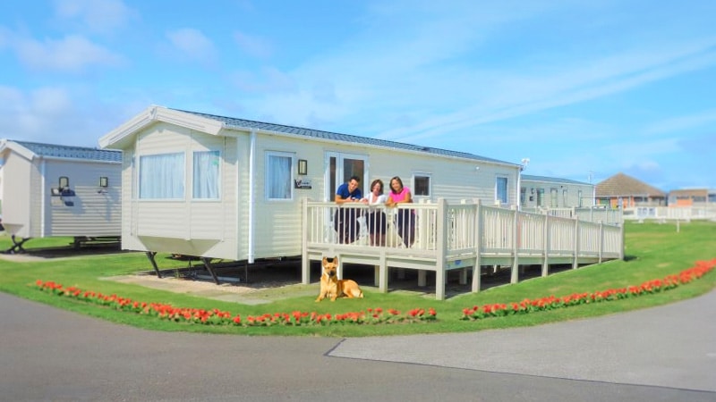 Cream-coloured dog friendly caravan holidays in ingoldmells, surrounded by vibrant red flowers, with a family enjoying the outdoor decking with their dog. Experience the charm of Kingfisher with this picturesque dog friendly caravan holidays in ingoldmells.