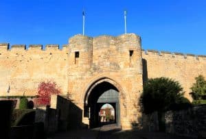Stone medieval gate in a fortified wall with an arched entrance and two towers, flags atop, under a clear blue sky.