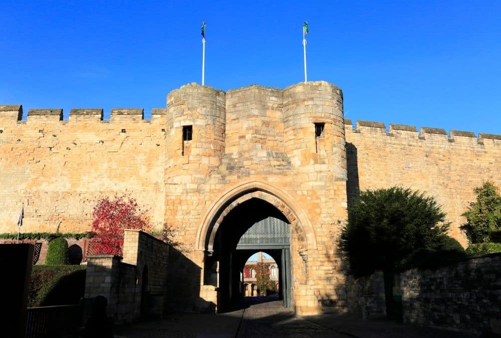 Stone medieval gate in a fortified wall with an arched entrance and two towers, flags atop, under a clear blue sky.
