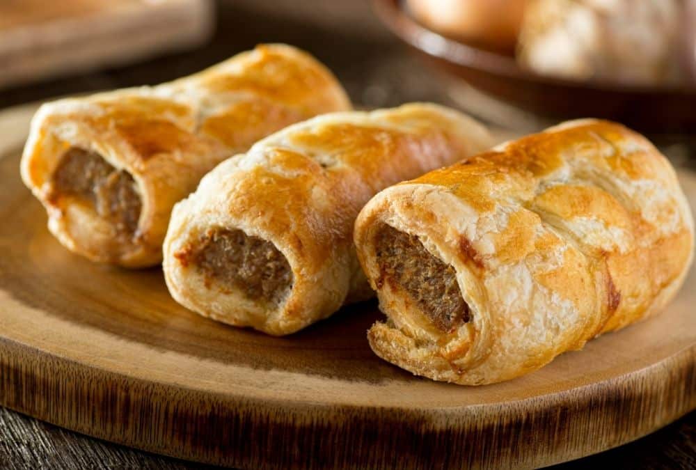 Three sausage rolls with golden flaky pastry on a wooden serving board, ends showing the sausage filling.