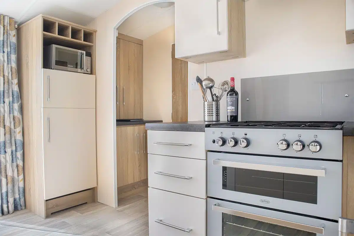 Fully equipped kitchen in Premium Plus 3 Bedroom Caravan featuring a full fridge-freezer, gas oven, and grill for all your cooking needs.