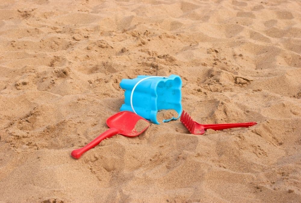 Blue plastic sand bucket overturned with red plastic shovel and rake on sandy beach.