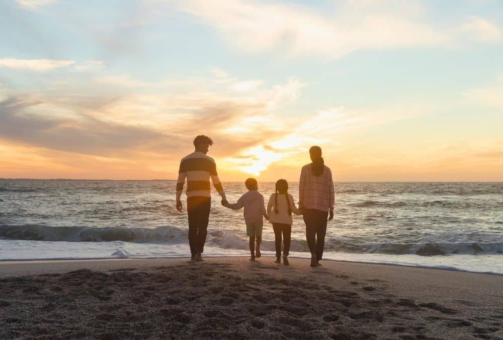 Family of four walking hand-in-hand along the beach at sunset.