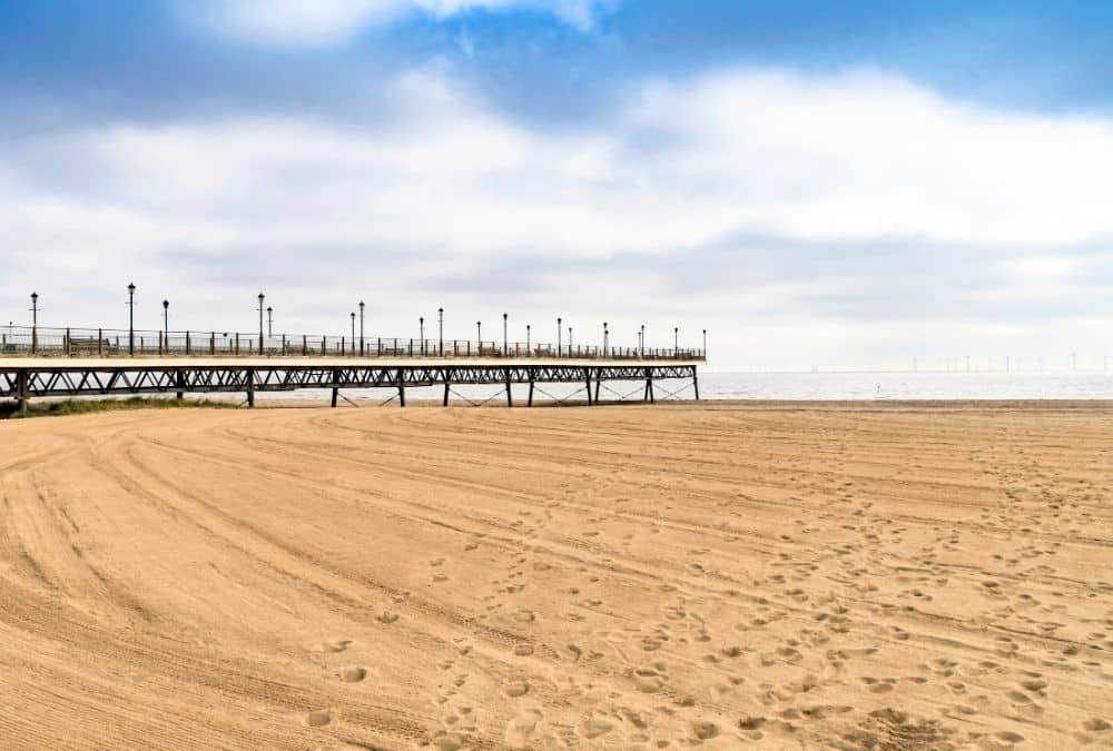 Pier extends into calm sea on a sandy beach under a partly cloudy blue sky with footprints in the sand nearby.