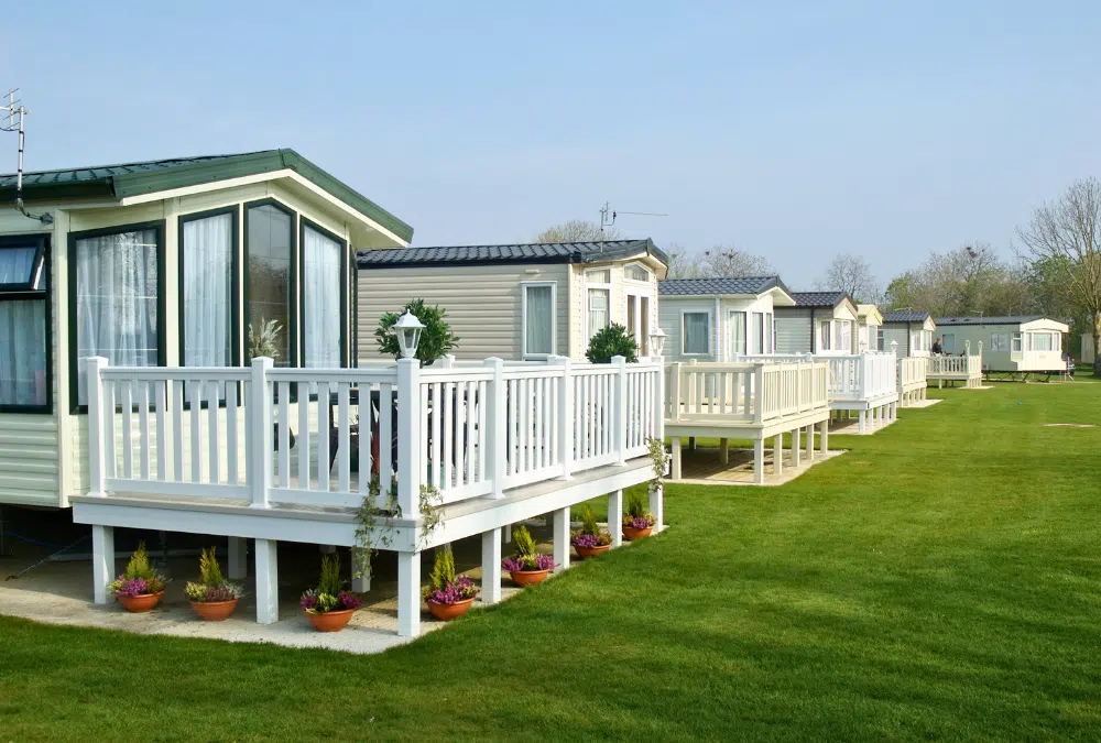 Row of white-railed porches on beige static caravans lined up along a grassy park, with potted plants and clear blue sky.