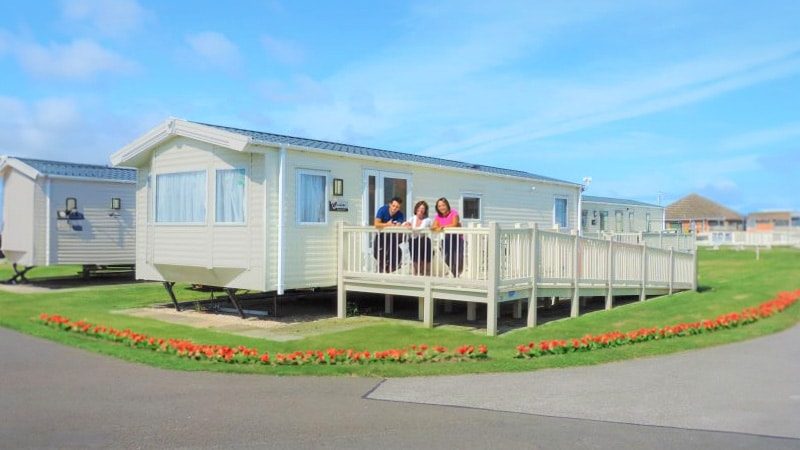 Cream-coloured caravan at Kingfisher Caravan Park, surrounded by vibrant red flowers, with a family enjoying the outdoor decking. Experience the charm of last minute caravans to rent in Skegness with this picturesque holiday scene